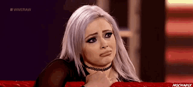 a woman with white hair and a choker is sitting on a red couch making a funny face .