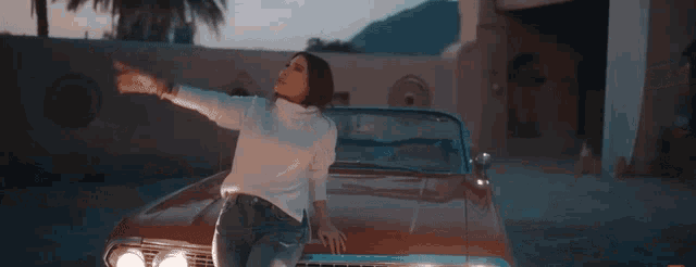 a woman in a white sweater is standing next to a red car with her arm in the air