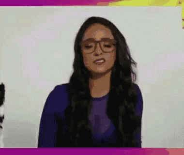 a woman wearing glasses and a purple shirt is standing in front of a white wall .