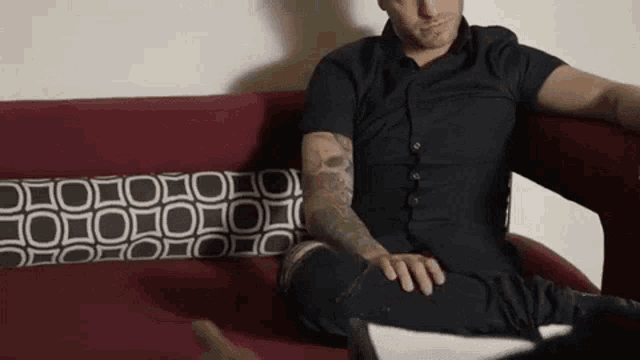 a man with tattoos is sitting on a red couch with a black and white pillow .