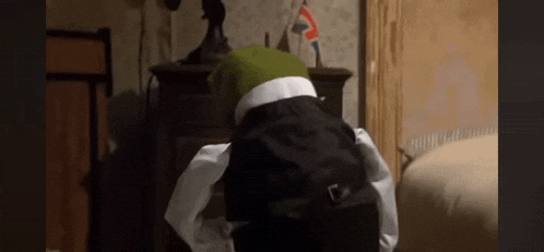 kermit the frog is wearing a black suit and white shirt and is kneeling down in a bedroom .