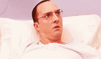 a man wearing glasses is laying in a hospital bed and making a surprised face .