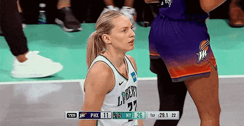 a woman in a ponytail is standing on a basketball court with a scoreboard behind her .