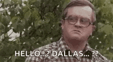 a man wearing glasses and a plaid shirt is standing in front of a tree and saying `` hello ... dallas ... '' .