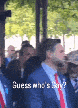 a man in a blue suit and red tie is standing in a crowd and says guess who 's gay
