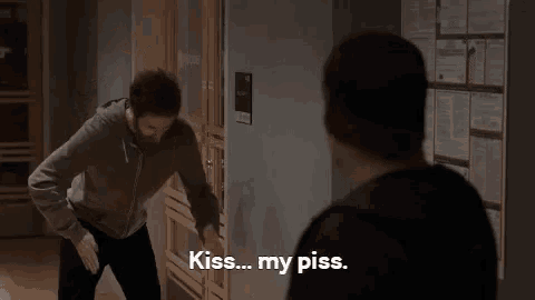 a man with a beard is standing next to another man in a hallway and says `` kiss ... my piss . ''