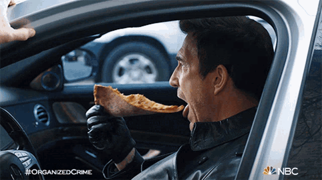 a man is eating a slice of pizza in a car with the nbc logo on the bottom right