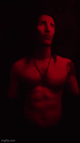 a shirtless man with long hair is standing in a dark room with a red light behind him .