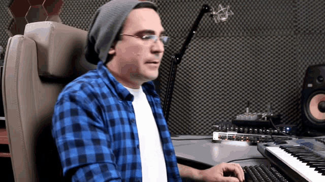 a man wearing glasses and a beanie is typing on a keyboard in front of a microphone