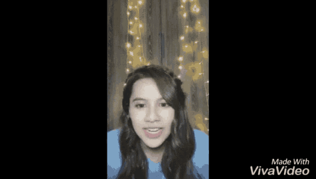 a woman in a blue shirt is standing in front of a wall with christmas lights behind her .