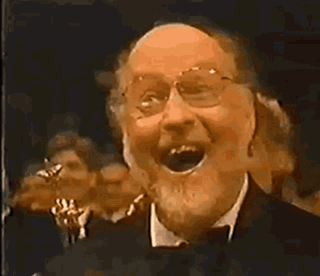 a man with glasses and a beard is laughing in front of a crowd of people .