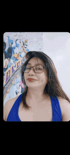 a woman wearing glasses and a blue tank top is smiling in front of a mickey mouse wall .