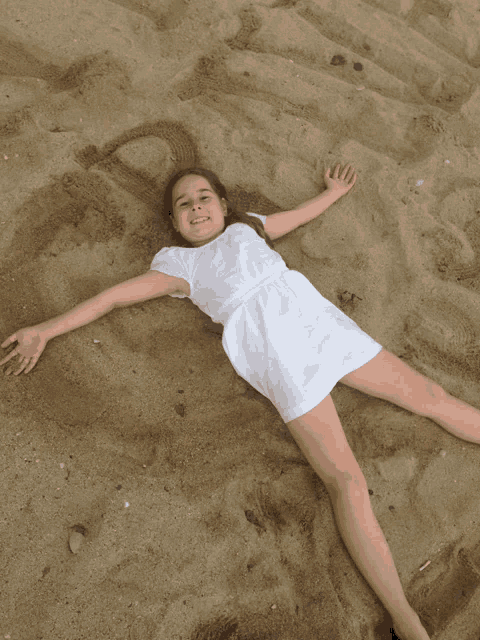 a young girl in a white dress is laying in the sand with her arms outstretched