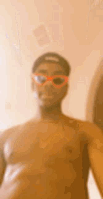 a shirtless man wearing sunglasses and a hat is taking a selfie in a bathroom .