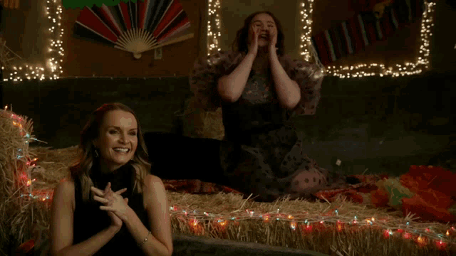 two women are sitting on hay bales with christmas lights