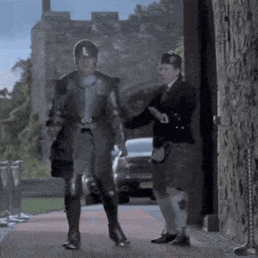 a man in armor and a man in a kilt are walking in front of a stone wall