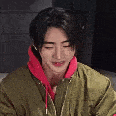 a young man wearing a green jacket and a red hoodie is smiling and looking at the camera .
