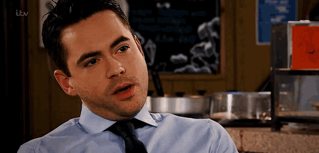 a man in a blue shirt and tie is sitting in front of a chalkboard that says itv on it