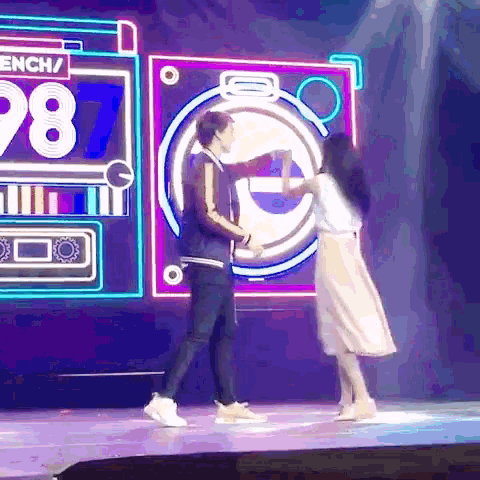 a man and woman are dancing on a stage in front of a neon sign that says 98
