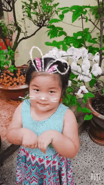 a little girl wearing a bunny ears headband stands in front of a plant with white flowers