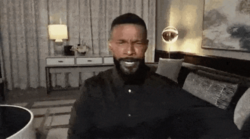 a man with a beard is sitting on a couch in a living room and smiling .