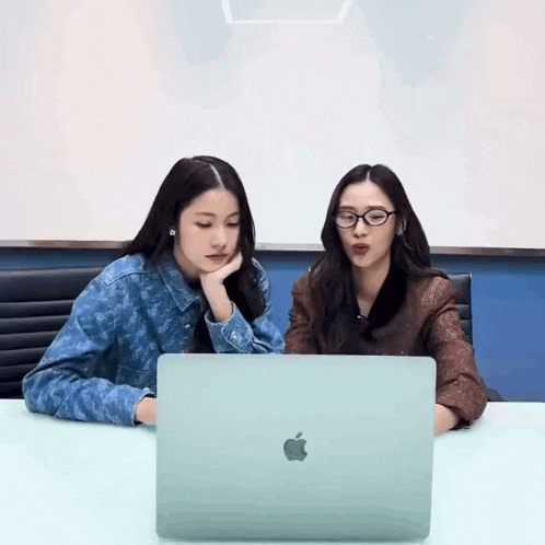 two young women are sitting at a table looking at an apple laptop .