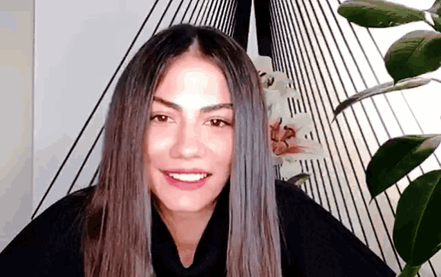 a woman with long hair is smiling and looking at the camera while standing in front of a plant .