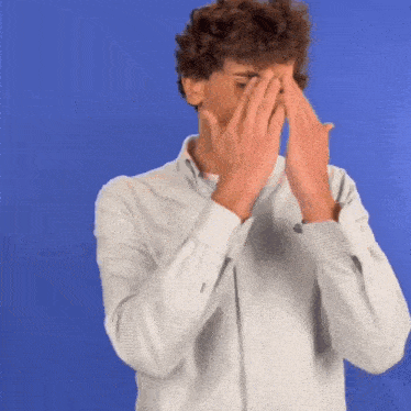 a man with curly hair is covering his face with his hands against a blue background .