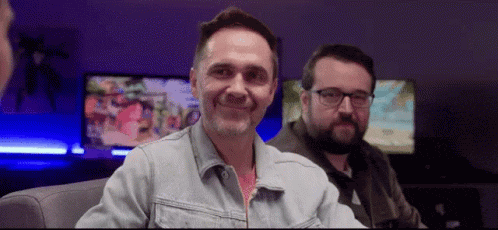 a man in a denim jacket is smiling while sitting next to another man in glasses .