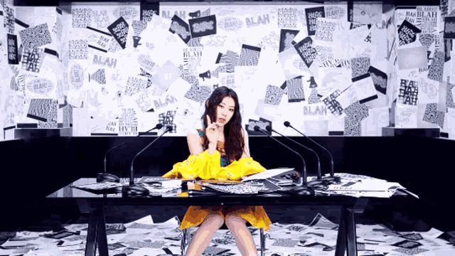 a woman in a yellow dress sits at a table in front of a wall that has blah written on it