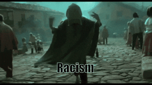 a man in a black cape is dancing on a cobblestone street with the word racism behind him