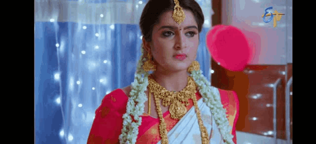 a woman in a red and white saree and gold jewelry is standing in front of a window .