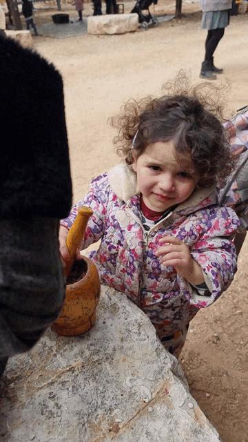 a little girl in a floral jacket is playing with a wooden mortar and pestle
