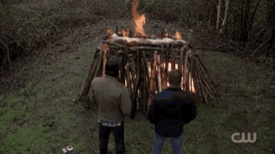 two men standing in front of a pile of logs with a cw logo on the bottom right
