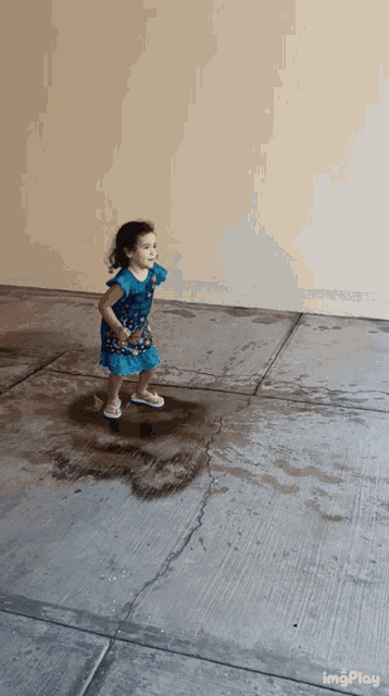 a little girl in a blue dress is jumping in a puddle of water on the sidewalk