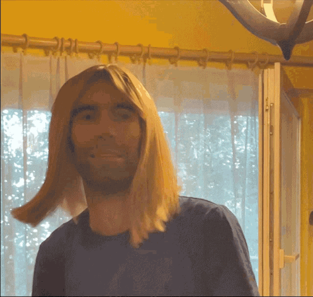 a man with long blonde hair is smiling in front of a yellow curtain
