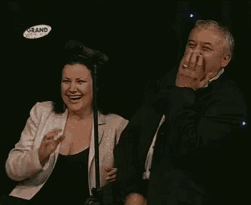 a man and a woman are laughing in front of a microphone with a grand logo in the corner