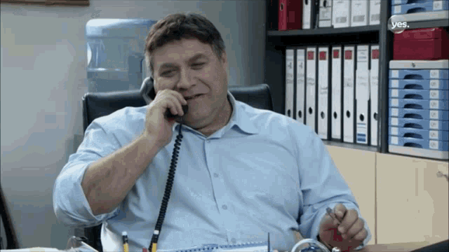 a man sitting at a desk talking on a phone with a yes logo on the bottom right