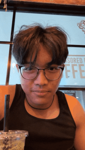 a young man wearing glasses and a black tank top is sitting in front of a sign that says sponsored coffee