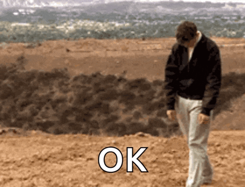 a man in a black jacket is walking in the dirt with the word ok written on the ground .