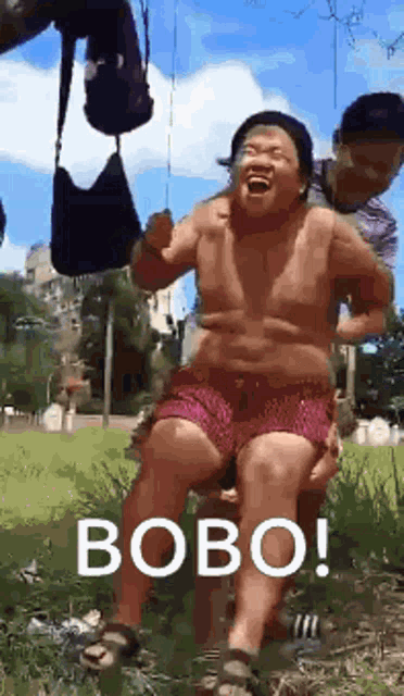 a shirtless man in red shorts is sitting on a swing with the word bobo written on it