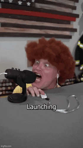 a man wearing a red wig is talking into a microphone with the word launching written below him