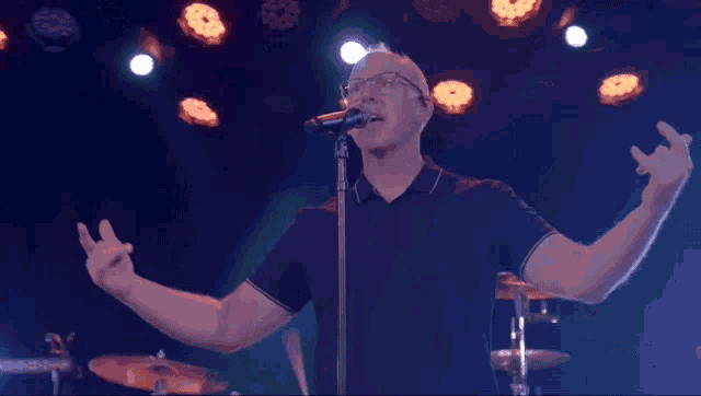 a man with glasses singing into a microphone with his arms outstretched
