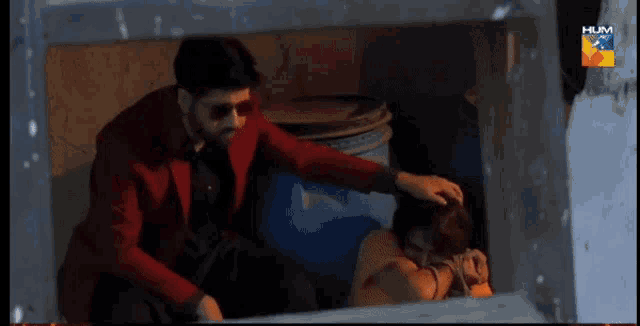 a man in a red jacket is putting his hand on a woman 's head who is tied up in a room on hum tv