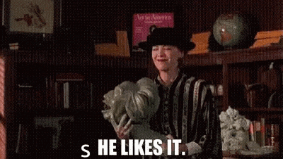 a woman in a hat is holding a vase of flowers in a library and says `` s he likes it '' .
