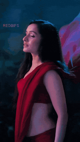 a woman in a red saree is standing in front of a purple flower .