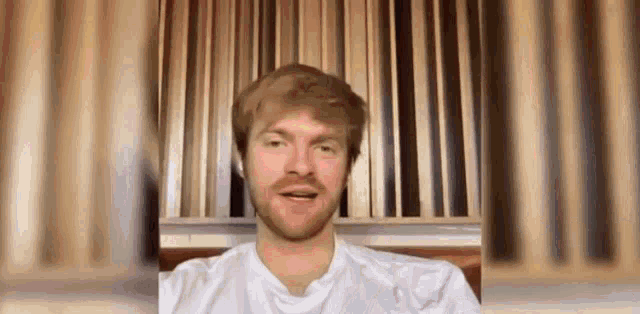 a man with a beard wearing a white shirt is sitting in front of a wooden wall .