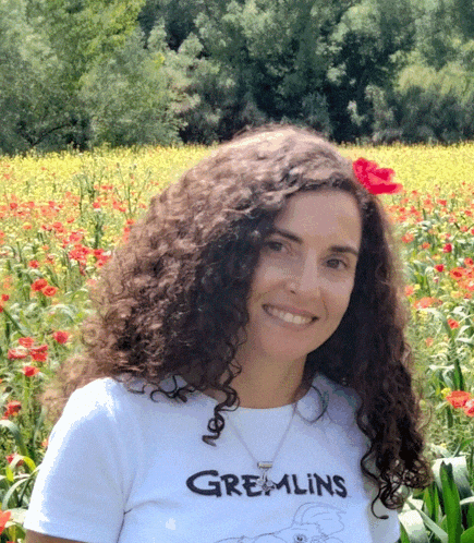 a woman wearing a gremlins shirt smiles in front of a field of yellow flowers