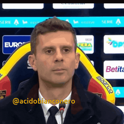 a man in a suit and tie stands in front of a microphone with the hashtag @acidobianconero on the bottom right