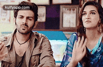 a man and a woman are sitting next to each other in a room and the woman is holding her hands to her chest .